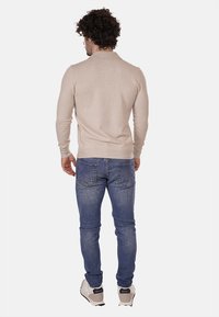 Maglione beige a maniche lunghe con colletto a girocollo, abbinato a jeans blu. La vista posteriore mostra il design aderente e il tessuto lavorato a maglia. Calzature casual.