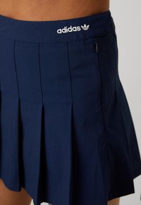 Navy plisirana suknja izrađena od lagane tkanine ima elastičnu trakicu u struku sa Adidas logotipom i bočni patentni zatvarač.