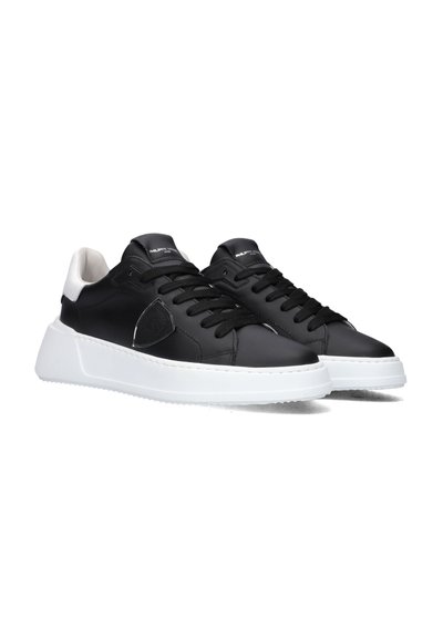 Black leather sneakers featuring a white rubber sole, round toe, and black laces. Includes a decorative patch on the side and a contrasting heel.