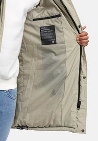 Interior de una chaqueta acolchada beige con textura suave, que presenta un bolsillo con cremallera y una etiqueta de cuidado con texto negro en una etiqueta.