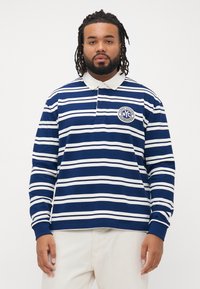 Mann med skjegg og dreads, iført en blå og hvitstripete langermet polo-skjorte med hvit krage og lyse bukser.