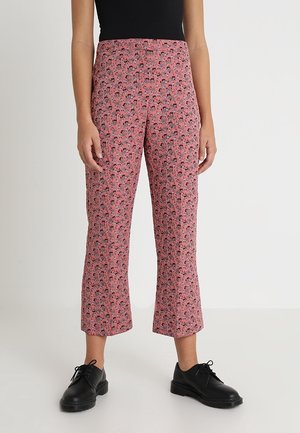 Persoon die een hoge taille roze bloemenkorte broek draagt met een zwart topje en zwarte veter schoenen, staand op een witte vloer en achtergrond.