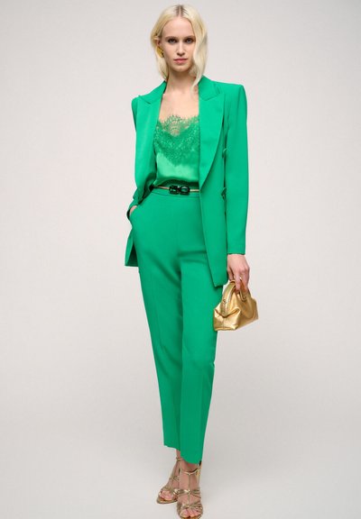 Costum verde ajustat cu o camiză din dantelă, talie accesorizată cu curea, accesorii din aur și sandale cu barete. Textură netedă și siluetă elegantă.