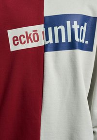 Panneaux de tissu rouges et blancs avec le texte de la marque « ecko » en rouge sur blanc et « unltd. » en blanc sur rectangles bleus, faisant partie d’un vêtement.