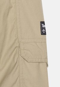 Tessuto beige con motivo a griglia, dotato di una tasca laterale e una patch nera con logo. Cuciture resistenti lungo i bordi.