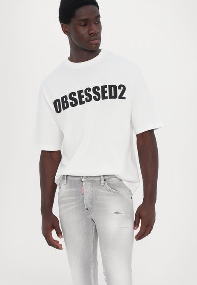T-shirt blanche à manches courtes avec "OBSESSED2" imprimé en lettres noires en gras. Porté avec un jean gris clair usé avec des déchirures subtiles.