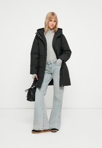 Čierna prešívaná parka s kapucňou, šedý dlhý rukáv, a svetlomodré široké nohavice. Čierna kabelka a topánky s hnedou podrážkou.