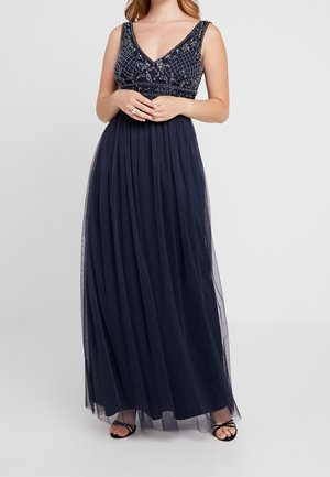 Femme portant une robe de soirée bleu marine sans manches avec un corsage orné de perles et une jupe fluide, debout les mains jointes à la taille.
