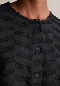 Schwarze Strickjacke mit strukturierten horizontalen Streifen, fransigen Kanten und runden schwarzen Knöpfen entlang der Frontöffnung. Weicher Stoff.