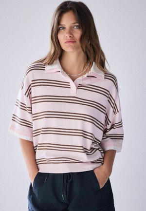 Frau mit langen braunen Haaren trägt ein lockeres rosa-braun gestreiftes Poloshirt und schwarze Hose, steht mit den Händen in den Taschen vor neutralem Hintergrund.