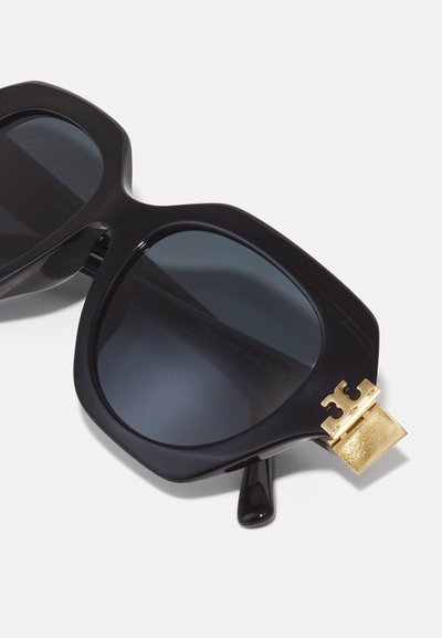 Black oversized sunglasses with dark lenses. Features a textured gold accent on the temple and distinctive thick frames.
