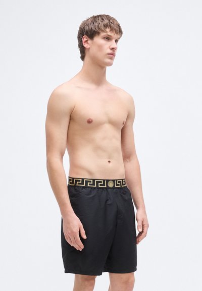 Zwarte korte broek met een gouden geruite tailleband en een gladde textuur. Het ontwerp is eenvoudig zonder extra patronen of accenten.