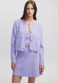 Lila Zweiteiler bestehend aus einer gekürzten Strickjacke mit goldenen Knöpfen und aufgesetzten Taschen, kombiniert mit einem passenden, ausgestellten Rock. Strukturierter Stoff.