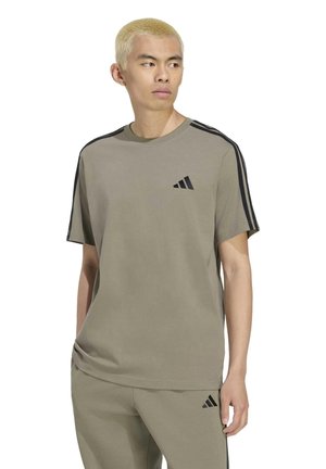 Modèle aux cheveux blonds courts portant un T-shirt Adidas couleur taupe et un pantalon assorti avec des détails à rayures noires, sur fond blanc.