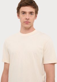 T-shirt color crema a maniche corte realizzato in tessuto morbido e leggero. Presenta un logo discreto nella parte superiore sinistra del petto.