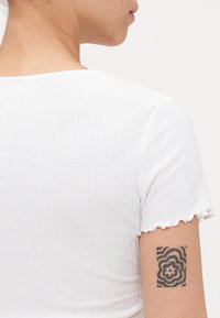 Biely rebrovaný top s krátkym rukávom a volánom na spodku, ktorý nosí osoba s vzorovaným tetovaním na hornej časti ramena.