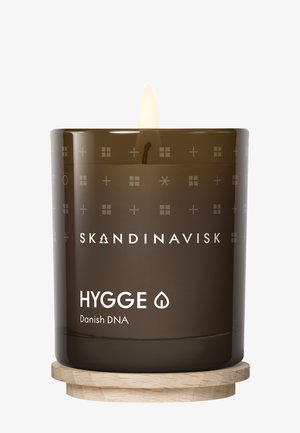 Bougie en verre marron avec une mèche allumée, portant le texte "SKANDINAVISK HYGGE" et des motifs géométriques décoratifs, posée sur un sous-verre en bois.