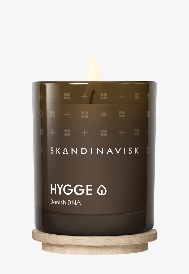 Bougie en verre marron avec une mèche allumée, portant le texte "SKANDINAVISK HYGGE" et des motifs géométriques décoratifs, posée sur un sous-verre en bois.