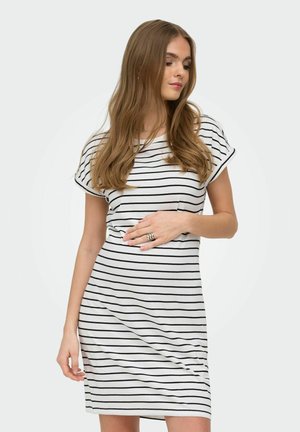 Kvinne med langt hår iført en hvit og svart kjole med horisontale striper, som forsiktig hviler hånden på magen og ser ned til siden.