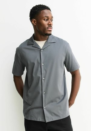 Homme portant une chemise grise à manches courtes boutonnée par-dessus un sous-vêtement blanc, regardant vers sa droite contre un fond blanc uni.