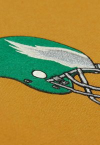 Grüner Footballhelm mit weißen Akzenten und grauem Gesichtsschutz auf einem senfgelben Stoffhintergrund bestickt.