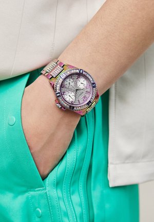 Poignet portant une montre métallique ornée de bijoux avec trois petits cadrans et des pierres étincelantes, main reposant dans une poche turquoise.