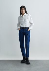 Λευκό πουκάμισο με κουμπιά και εφαρμοστή μέση, μπλε τζιν παντελόνι, μαύρη ζώνη και χοντρά μαύρα παπούτσια. Στέκεται μπροστά σε ουδέτερο φόντο.