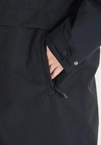 Svart jacka med en dragkedjeficka och tryckknappar. Tyget har en slät yta med subtila diagonala mönster.