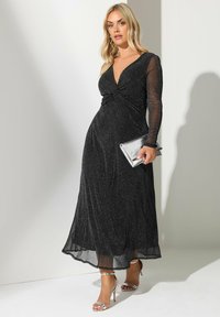 Musta maxi-mekko, jossa on kimaltava pinta, syvä V-pääntie, pitkät läpinäkyvät hihat ja istuva vyötärö. Yhdistettynä metallinhohtoiseen clutchiin ja hopeisiin korkokenkiin.