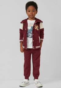 Bordo sportinis kostiumas, kuriame yra sukapotas džemperis su baltomis detalėmis, grafiškas marškinėliai ir derančios kelnės su juostelėmis, papildytas baltomis sportinėmis bateliais.