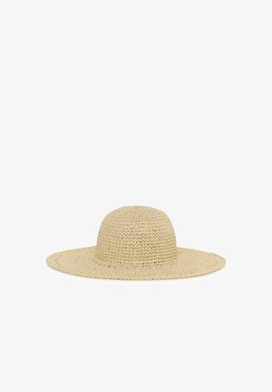 Vrouw die een mouwloze beige jurk met verticale grijze strepen draagt en een breedgerande geweven zonnehoed, staand met handen in de zakken.