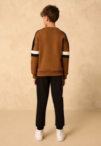 Sweat-shirt marron avec des accents rayés noirs et blancs, associé à un pantalon noir. Le modèle porte des baskets blanches. Fond neutre.