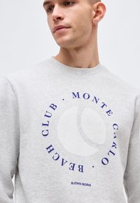 Άνδρας που φοράει ανοιχτό γκρι φούτερ με μπλε κυκλικό κείμενο «Monte Carlo Beach Club» και μια γραφική απεικόνιση μπάλας τένις στο κέντρο.