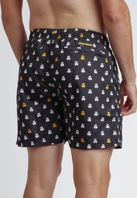 Bañador negro con un patrón repetitivo de caras de dibujos animados en blanco y amarillo, que cuenta con una cintura elástica y un sutil bolsillo lateral.