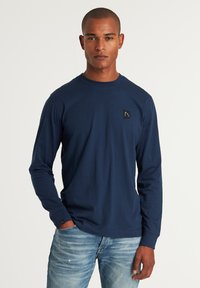 Marineblaue Langarmbluse aus weichem Baumwollmaterial mit Rundhalsausschnitt und einem kleinen schwarzen Logopatch auf der linken Brust. Mit enganliegenden Bündchen.