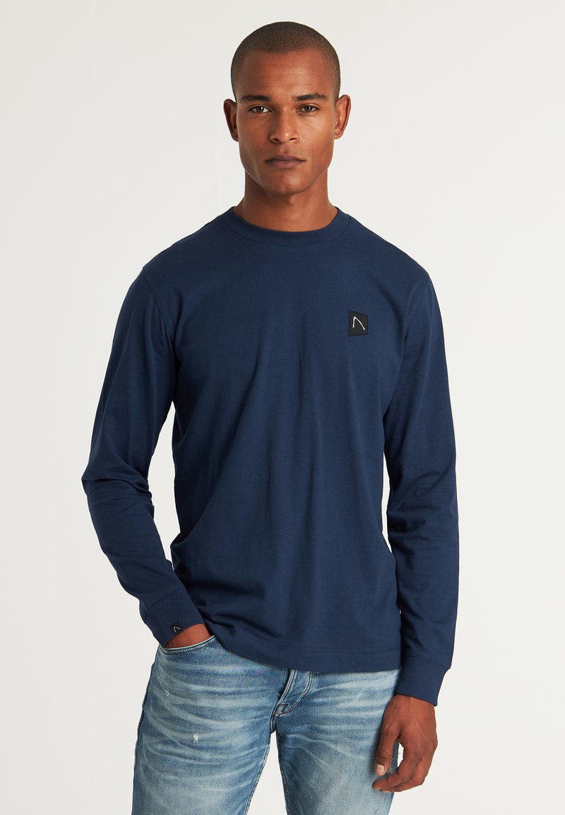 Marineblaue Langarmbluse aus weichem Baumwollmaterial mit Rundhalsausschnitt und einem kleinen schwarzen Logopatch auf der linken Brust. Mit enganliegenden Bündchen.
