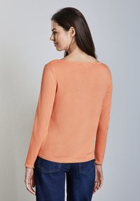 Sweter z długim rękawem w koralowym kolorze, z szerokim dekoltem i ściągaczami przy mankietach. W zestawie z niebieskimi dżinsami. Prosty, dopasowany krój.