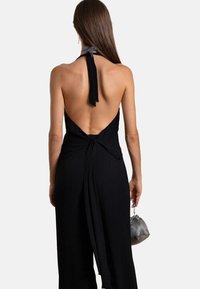 Rückansicht eines schwarzen Jumpsuits mit Halter, offener Rückseite, Bindeband am Ausschnitt und fließendem Stoff; kombiniert mit einer kleinen silbernen Tasche.