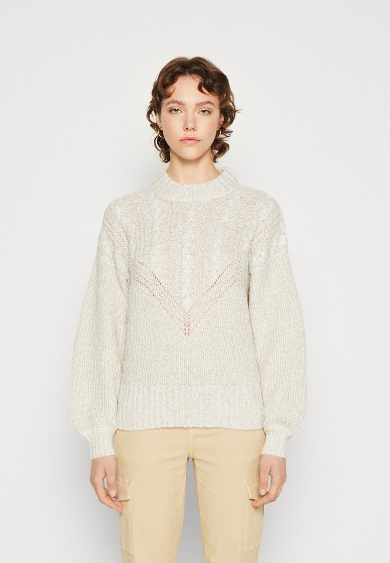 Lys beige strikket sweater med høj hals, tekstureret mønster og ballonærmer. Har et V-formet design på forsiden.