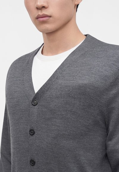 Homme portant un cardigan gris foncé boutonné par-dessus un t-shirt blanc à col rond, montrant le haut du torse et une partie du visage sur un fond uni.