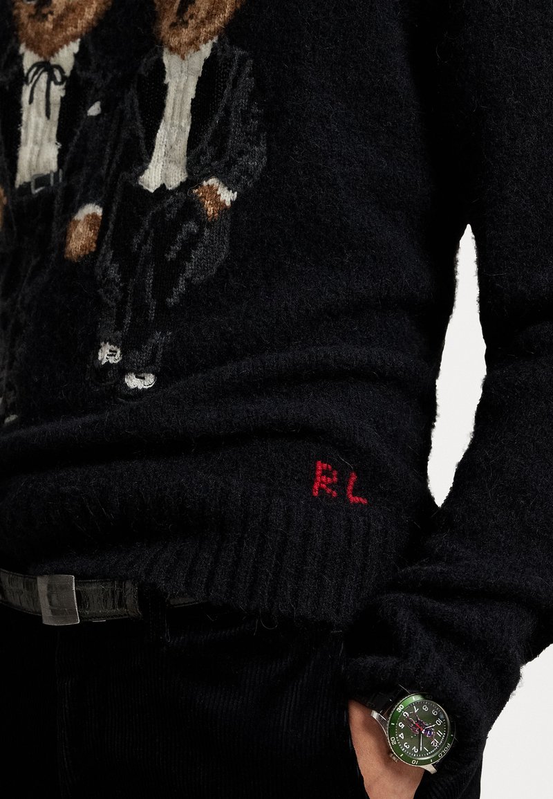 Pull en maille noir avec deux ours en smoking. La manche gauche arbore une broderie rouge « RL ». Une montre verte est portée au poignet.