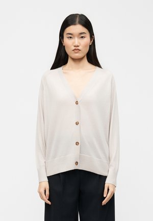 Hosszú fekete hajú nő világos bézs, gombos kardigánban és fekete nadrágban, egyszerű fehér háttér előtt állva.