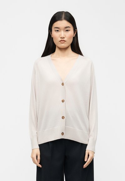 Femme aux longs cheveux noirs portant un cardigan beige clair boutonné et un pantalon noir, debout devant un fond blanc uni.