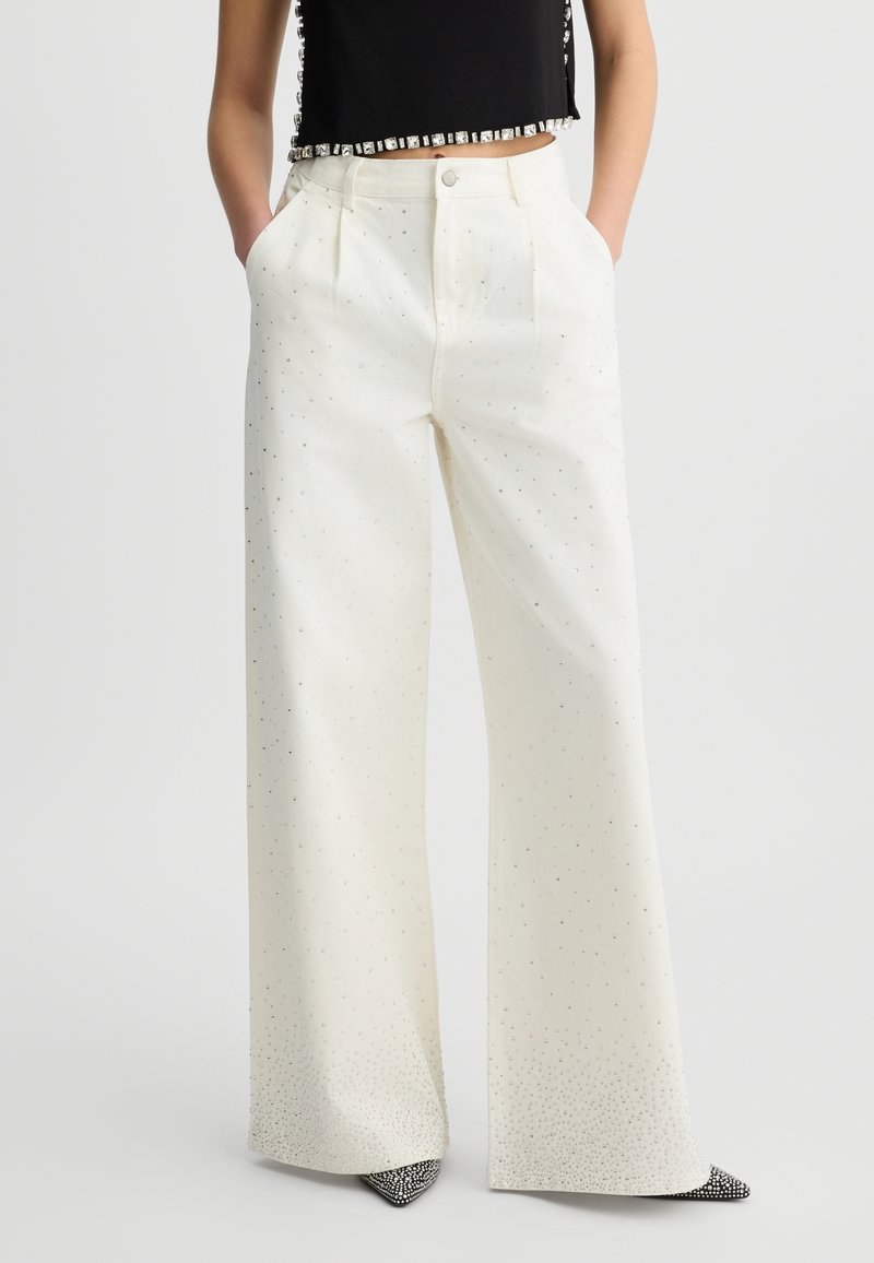 Pantalons larges en tissu crème, avec un motif subtil de petits accents argentés, des poches avant et une fermeture à bouton.