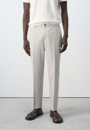 Mann, der beige plissierte Hosen, ein weißes kurzärmeliges Oberteil und schwarze Sandalen trägt und vor einem einfarbigen hellen Hintergrund steht.