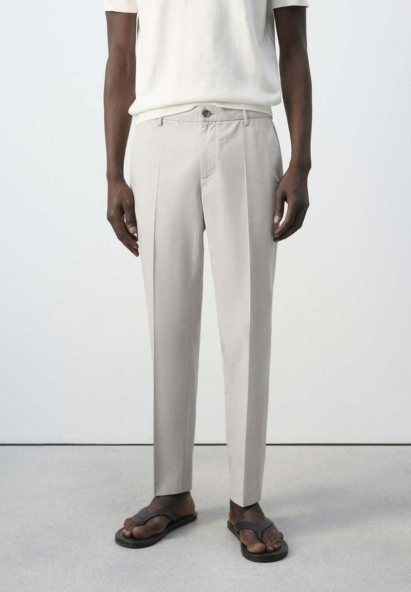 Homme portant un pantalon plissé beige, un haut blanc à manches courtes et des sandales noires, debout devant un fond clair uni.