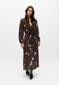 Mujer de pie que lleva un vestido cruzado marrón con estampados florales en azul claro, cinturón atado, mangas largas y botines negros puntiagudos.