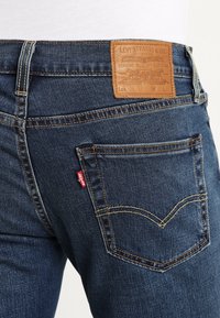 Granatowe dżinsy z denimu z skórzaną metką na pasku, z pomarańczowymi szwami i czerwoną etykietą na tylnej kieszeni.