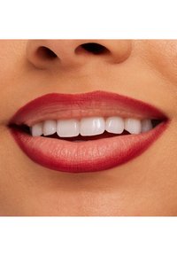 Zbliżenie na usta z głęboko czerwonym matowym pomadką, gładką teksturą, widocznymi białymi zębami oraz naturalnym kolorem skóry w tle.