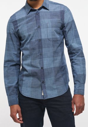 Chemise à carreaux bleus à manches longues, avec une poche avant et un tissu texturé. Se distingue par des boutons noirs et un ourlet arrondi.